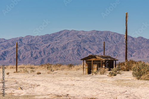 USA, CA, Salton Sea - December 28, 2012: Wooden brown shack lost in dunes of beige sand with greenish brown shrubs under blue sky. Brown mountain range on horizon.