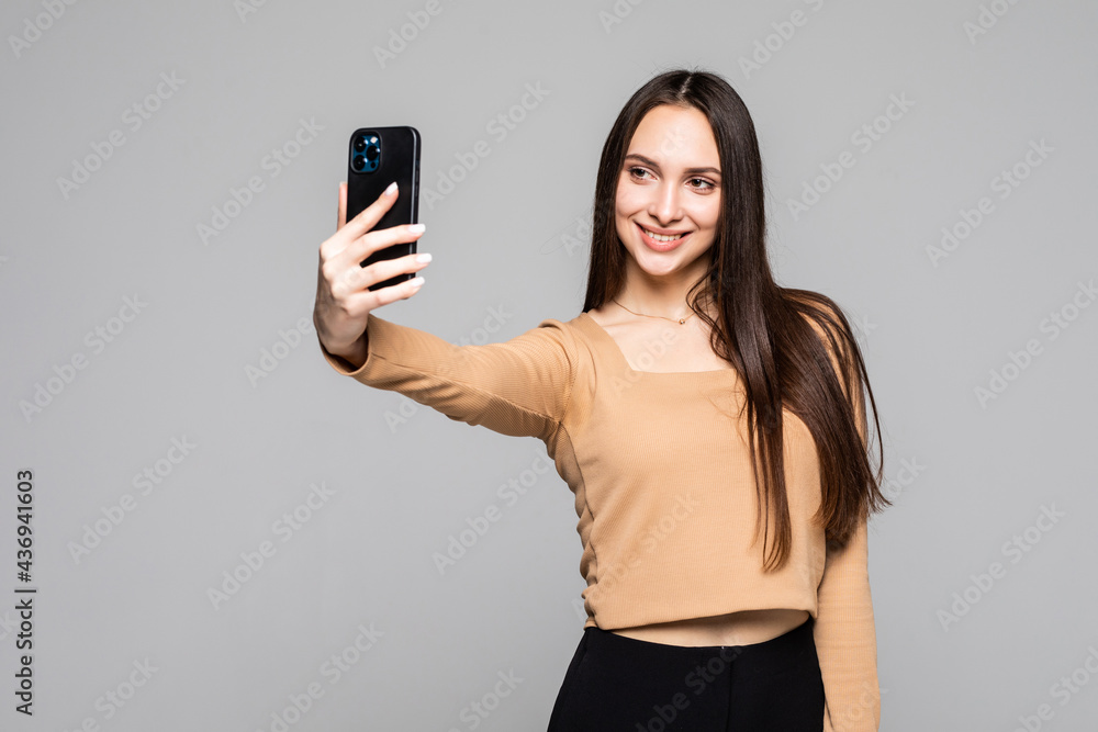 Naklejka premium Smiling young woman making selfie photo on smartphone over gray background