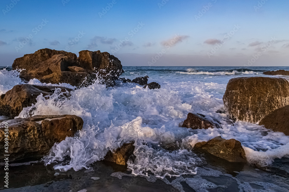 Closeup of foamy wave rushing ashore amid rockly coast of Laguna Beach, California. Early morning sky, clouds in the distance. 

