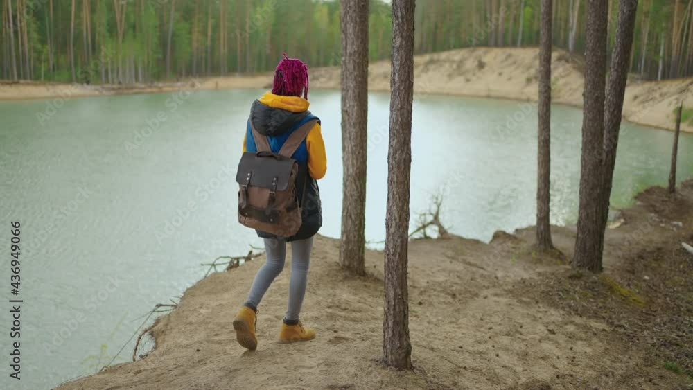 A young black woman in a yellow cursor climbs up with a backpack uphill in the woods looking at the lake. Slow motion A young black woman in a yellow cursor climbs up with a backpack uphill in the woods looking at the lake. Slow motion