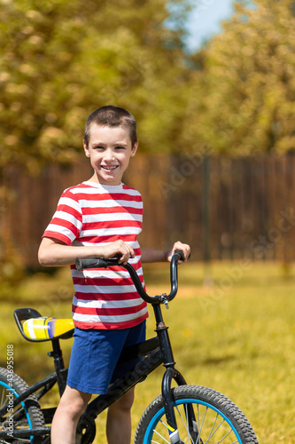 Happy cheerful boy in a striped t-shirt is doing well, looking at the camera and sitting on a bike against the background of the garden. Active outdoor games for children in summer.