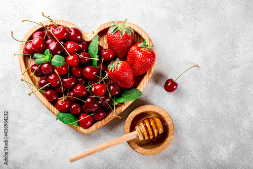 Ripe cherries and strawberries on a wooden plate in the shape of a heart and honey on a light gray background