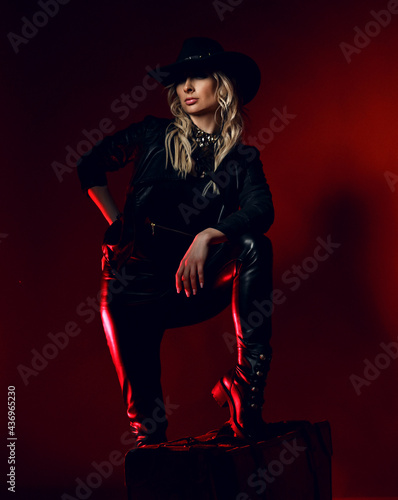 Cool sexy curly blonde woman cowgirl in black leather pants and wide-brimmed stetson hat stands with her foot on chair, leaning at her knee over dark red background.