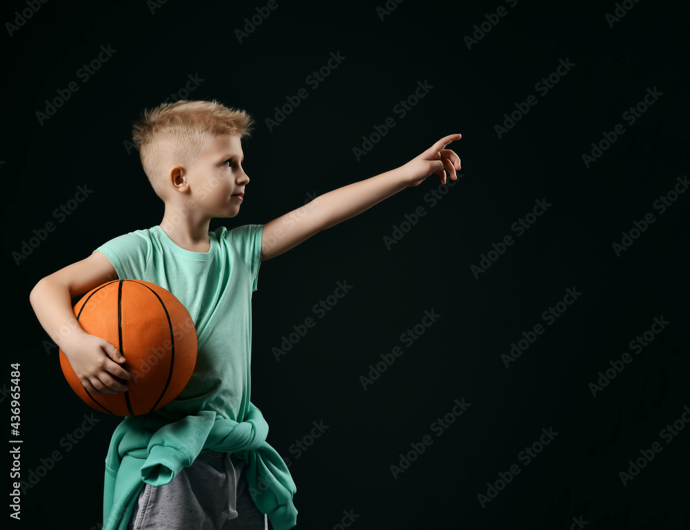 Smiling blond boy child in sports green t-shirt and trousers standing holding basketball in hands pointing up with finger over dark background. Trendy sports active children fashion concept