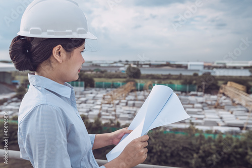 Female Civil Engineer in Safety Hard Hat With Blueprint Paper While Control Production in Manufacturing Factory. Portrait of Professional Construction Engineer During Working at Building Project Site.