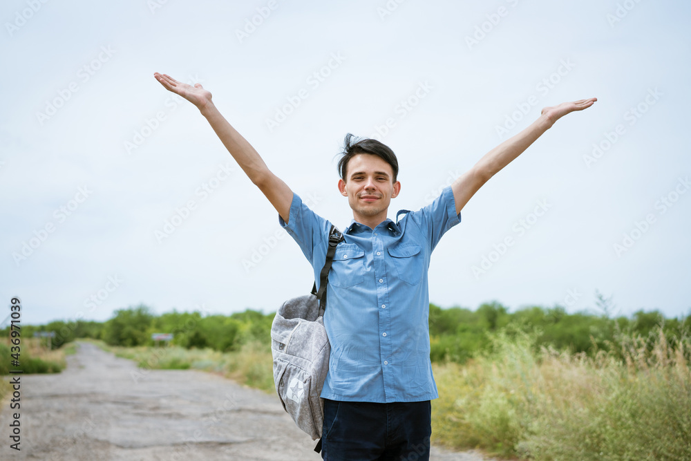 Happy young man posing with raised arms, standing on the road and looking into the distance. Happy guy in a blue shirt with a backpack. Free student enjoys vacation Happy young man posing with raised arms, standing on the road and looking into the distance. Happy guy in a blue shirt with a backpack. Free student enjoys vacation