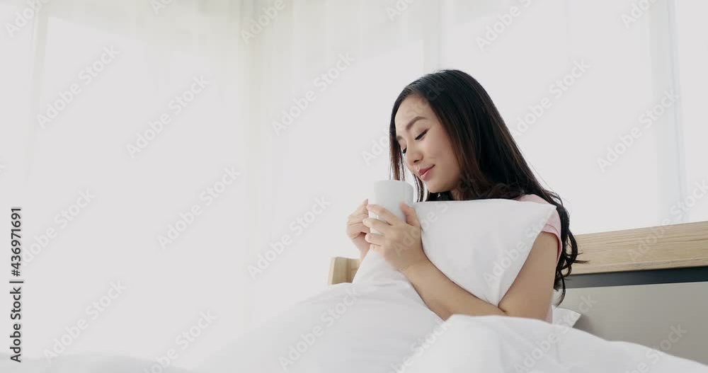 Young beautiful attractive asian women with long hair sitting on her bed in the morning sipping coffee while hugging her white pillow. Bright background