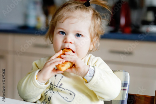 Happy little baby girl eating fresh croissant for breakfast or lunch. Healthy eating for children. Toddler child in colorful pajama sitting in domestic kitchen after sleeping in the morning 