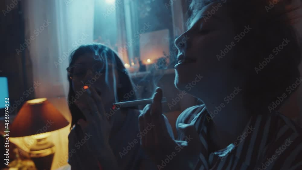 Two young girlfriends lighting cigarettes and smoking in dark room ...