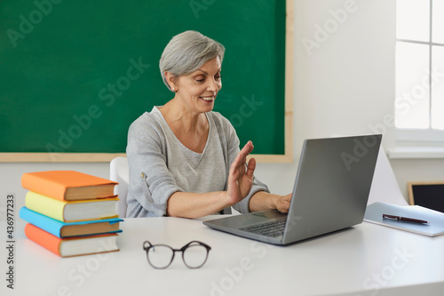 Online schooling. Senior teacher waving to her students while communicating on laptop at classroom. Mature tutor giving lesson or web conference