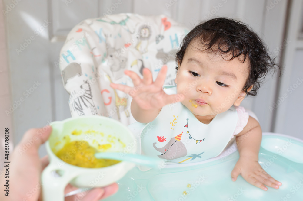 A hungry Asian baby girl sitting at the dining table looking at the food on the plate in her mother hand