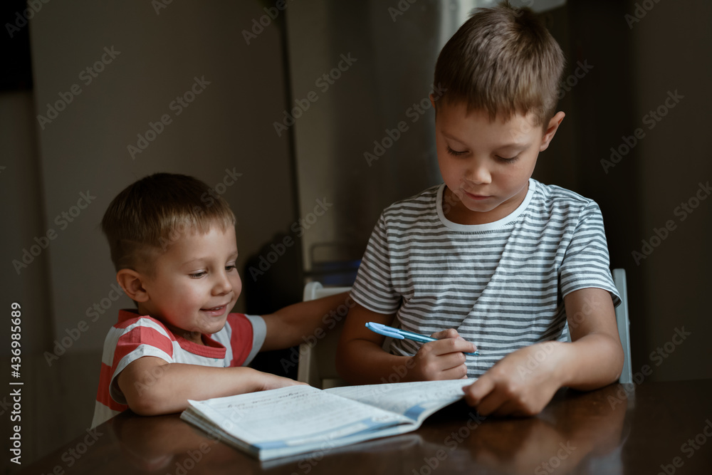 cute caucasian boy writing letters in a copy book. His little brother looking at him. Image with selective focus cute caucasian boy writing letters in a copy book. His little brother looking at him. Image with selective focus