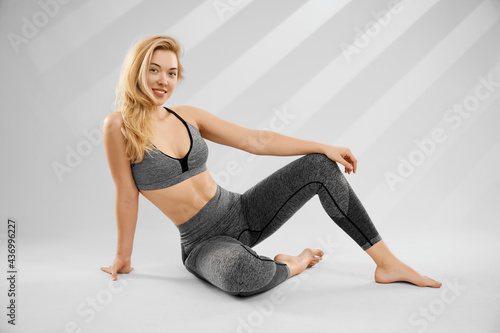 Front view of smiling young blonde woman in trendy sportswear sitting on grey background in photo studio. Concept of preparing for doing meditation or yoga for improve well-being. 