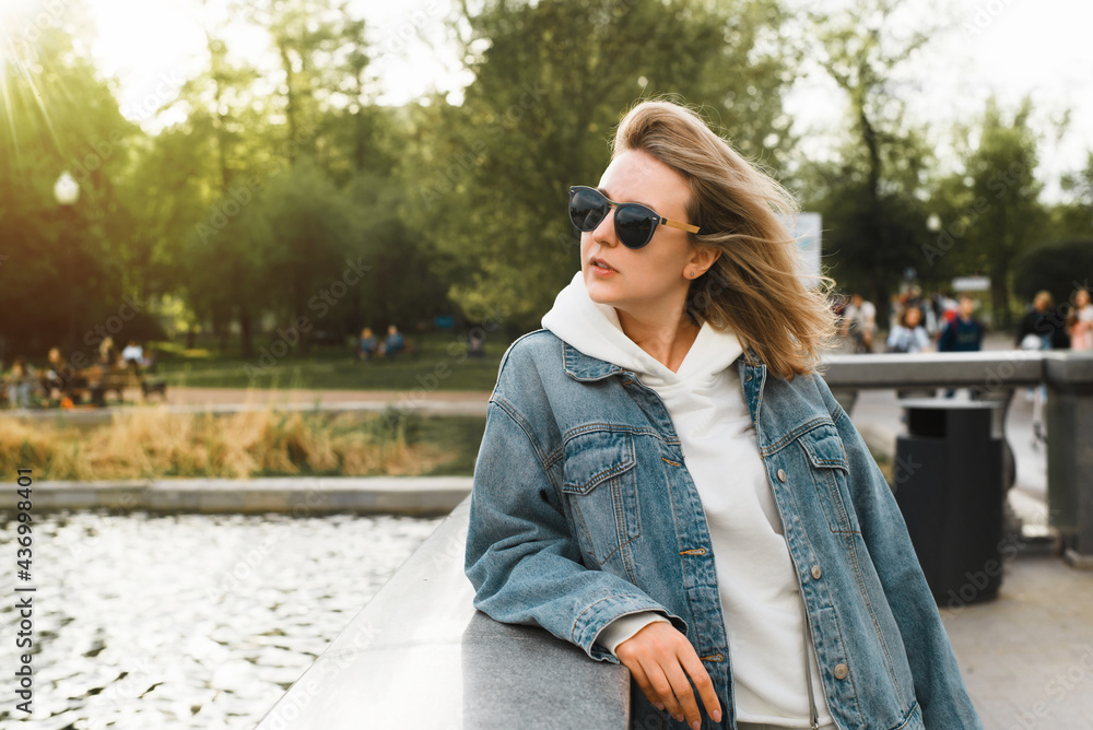 Stylish female portrait lifestyle. Woman in glasses and jeans standing on the bridge and looking to the side in a city park, profile photo. Blonde girl enjoying a weekend walk in nature.
