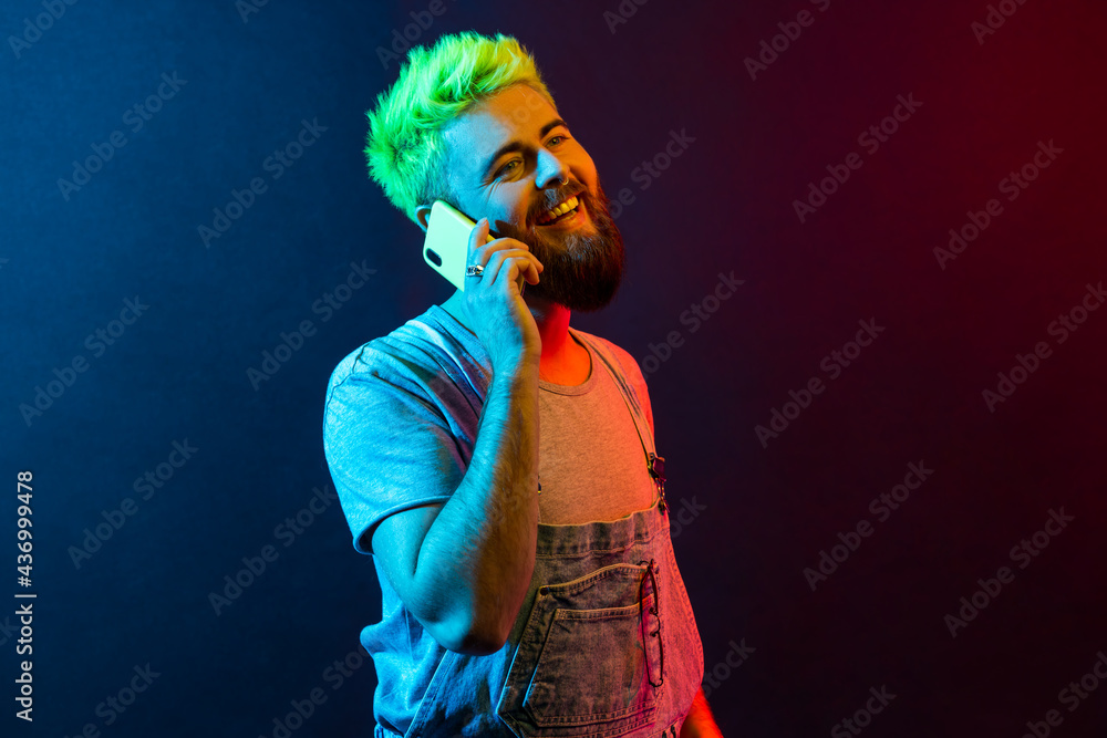 Portrait of young adult hipster man wearing denim overalls and t shirt talking phone, hears happy news, smiles toothily, expressing optimism. Colorful neon light, indoor studio shot.