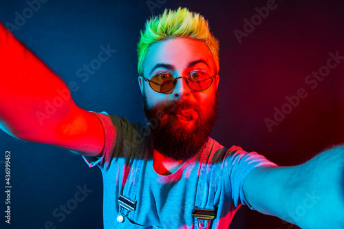Extremely crazy bearded hipster man blogger with green hair wears denim overalls, showing tongue out, having fun posing on selfie camera, live streaming. Colorful neon light, indoor studio shot.