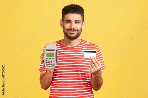 Smiling man with beard in striped t-shirt holding and showing pos payment terminal and credit or debit card, using cashless payments, nfc. Indoor studio shot isolated on yellow background © khosrork