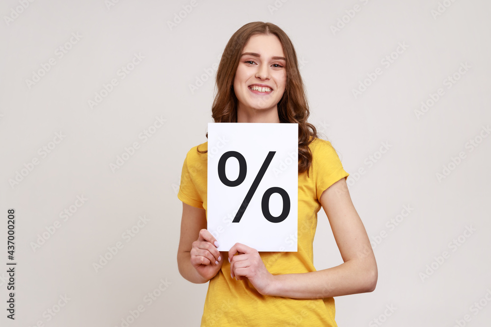 Smiling smart girl with brown wavy hair wearing yellow casual T-shirt holding paper and shows with percent sign inscription, looking at camera. Indoor studio shot isolated on gray background. Smiling smart girl with brown wavy hair wearing yellow casual T-shirt holding paper and shows with percent sign inscription, looking at camera. Indoor studio shot isolated on gray background.