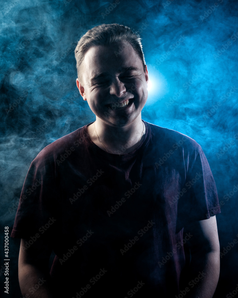 The guy laughs and smiles against the background of smoke, consecrated in blue