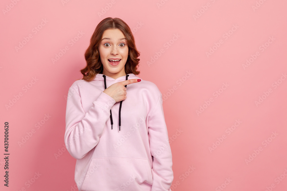 Portrait of surprised young female with curly hair in hoodie looking at camera with happy amazed expression, pointing finger right at copy space. Indoor studio shot isolated on pink background Portrait of surprised young female with curly hair in hoodie looking at camera with happy amazed expression, pointing finger right at copy space. Indoor studio shot isolated on pink background