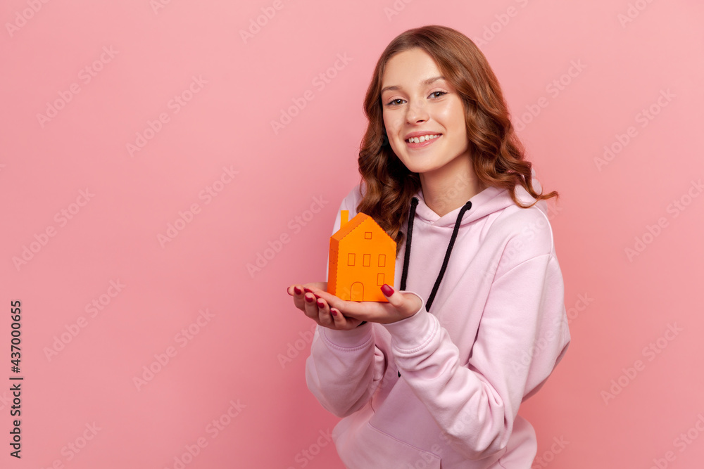Portrait of happy teen girl in hoodie holding in hands little paper house looking at camera with smile, dreaming about her own apartment. Indoor studio shot isolated on pink background Portrait of happy teen girl in hoodie holding in hands little paper house looking at camera with smile, dreaming about her own apartment. Indoor studio shot isolated on pink background