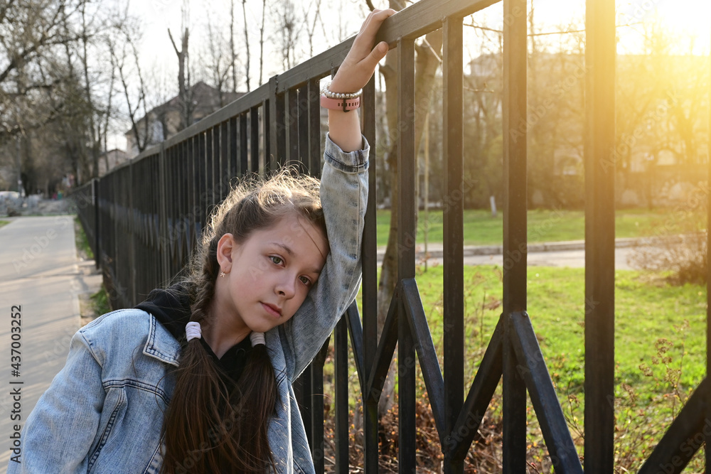 pretty, thoughtful girl in denim clothes is standing by a metal fence, leaning against it and looking into the distance. Close-up portrait in the sunlight