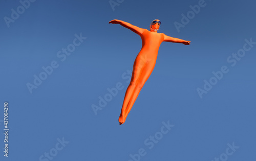 A Girl is dressed in an orange bodysuit. She wears a flying cap with flying goggles. She jumps up high into mid-air and pretends to be an airplane.