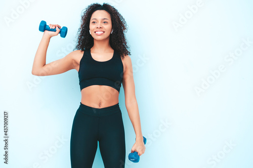 Fitness smiling black woman in sports clothing with afro curls hairstyle.She wearing sportswear. Young beautiful model with perfect tanned body.Female holding dumbbells in studio near light blue wall