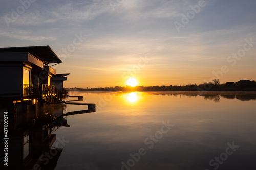 Beautiful orange sunrise over the river. Golden twilight, sunlight reflected in water ripples. Dark silhouette of the trees, rays of sunset reflected in the windows. Vacation, tranquility. © Maya_parf