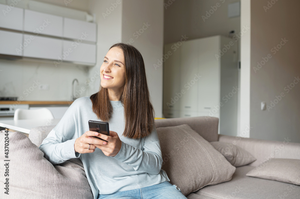 Cheerful dreaming woman holds smartphone sitting on the couch at home, thoughtfully looks aside, chatting online with friends, spends leisure time in social media Cheerful dreaming woman holds smartphone sitting on the couch at home, thoughtfully looks aside, chatting online with friends, spends leisure time in social media