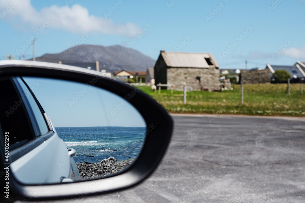 Obraz premium Blue ocean surface reflection in a car mirror in focus. Small town and a mountain out of focus in the background. Travel and tourism concept. Achill island, county Mayo, Ireland. Warm day