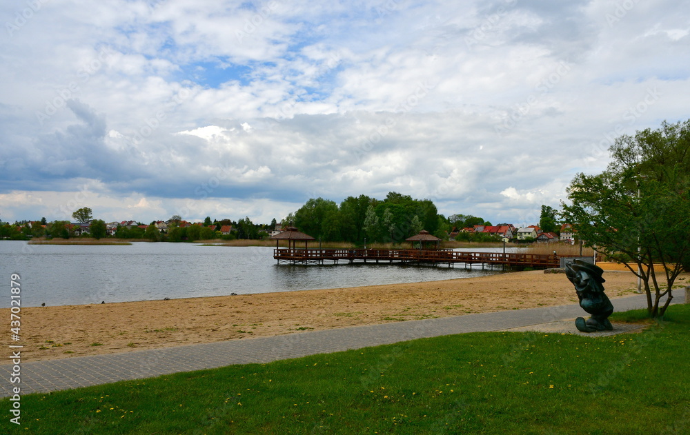 A view of a small sandy beach located next to the edge of a shallow yet vast river or lake situated next to a well maintained public park with some pier or marina visible in the background in Poland