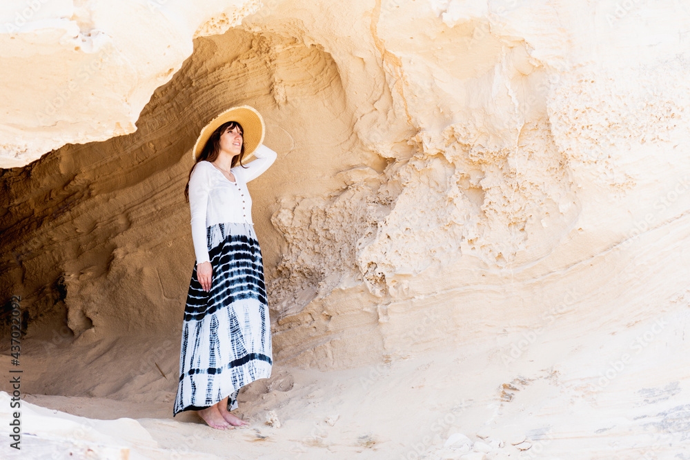 Naklejka premium woman in a cave on the beach looking at the sea, wears long dress and straw hat