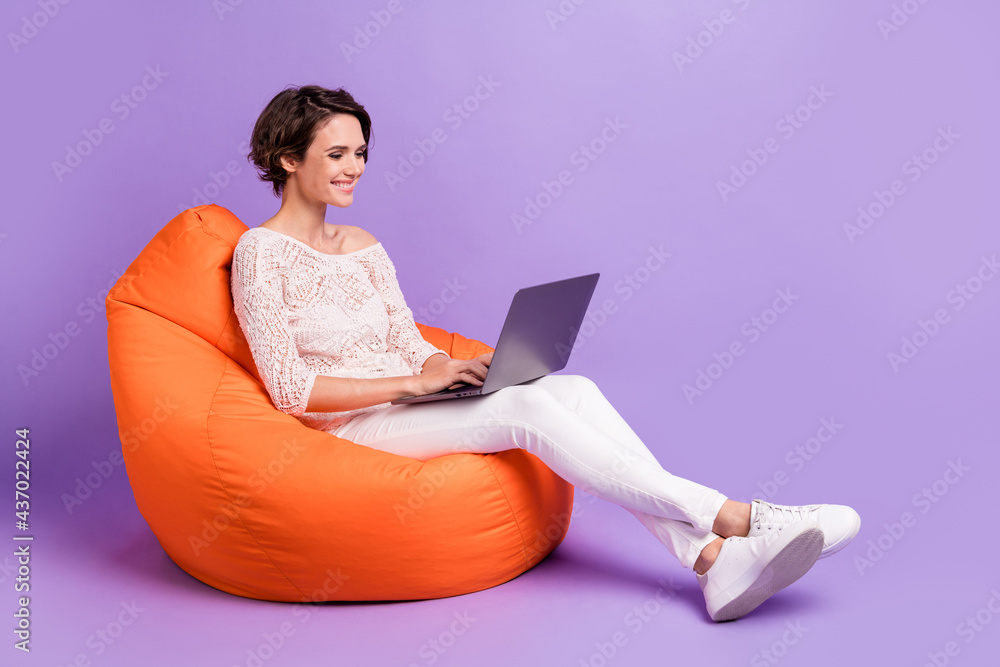 Full length profile photo of charming cheerful lady sit big soft chair use laptop isolated on violet color background Full length profile photo of charming cheerful lady sit big soft chair use laptop isolated on violet color background