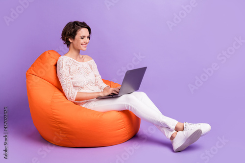 Full length profile photo of charming cheerful lady sit big soft chair use laptop isolated on violet color background