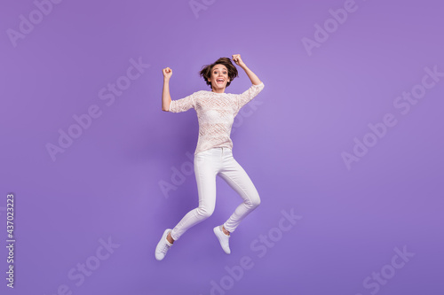 Full length photo of funny lucky young woman wear knitted shirt jumping high rising fists isolated violet color background