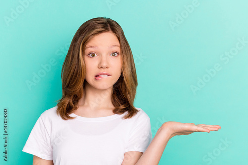 Photo portrait little girl biting lip keeping blank space on hand isolated vivid turquoise color background