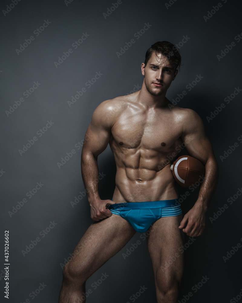 Shirtless man with american football ball in blue underwear at grey background in studio Shirtless man with american football ball in blue underwear at grey background in studio