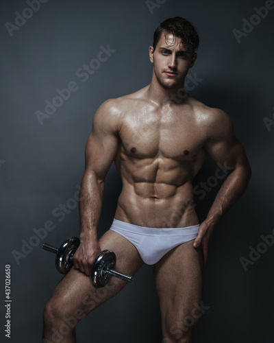 Muscular young man in white underwear with dumbbell in studio at grey background