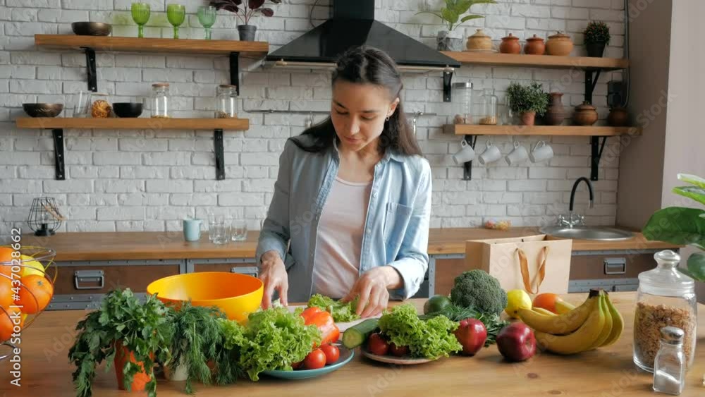 Happy young housewife prepares vegetable salad in her kitchen.Beautiful young woman in a good mood cuts lettuce leaves for salad. Young beautiful woman cuts vegetables for salad and puts them in a Happy young housewife prepares vegetable salad in her kitchen.Beautiful young woman in a good mood cuts lettuce leaves for salad. Young beautiful woman cuts vegetables for salad and puts them in a