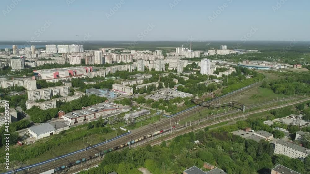 Aerial view of freight train leaving town. Barrel train. High and low residential buildings nearby and industrial zone. There are many trees in the city. Endless forests on the horizon. Sunny summer d Aerial view of freight train leaving town. Barrel train. High and low residential buildings nearby and industrial zone. There are many trees in the city. Endless forests on the horizon. Sunny summer d