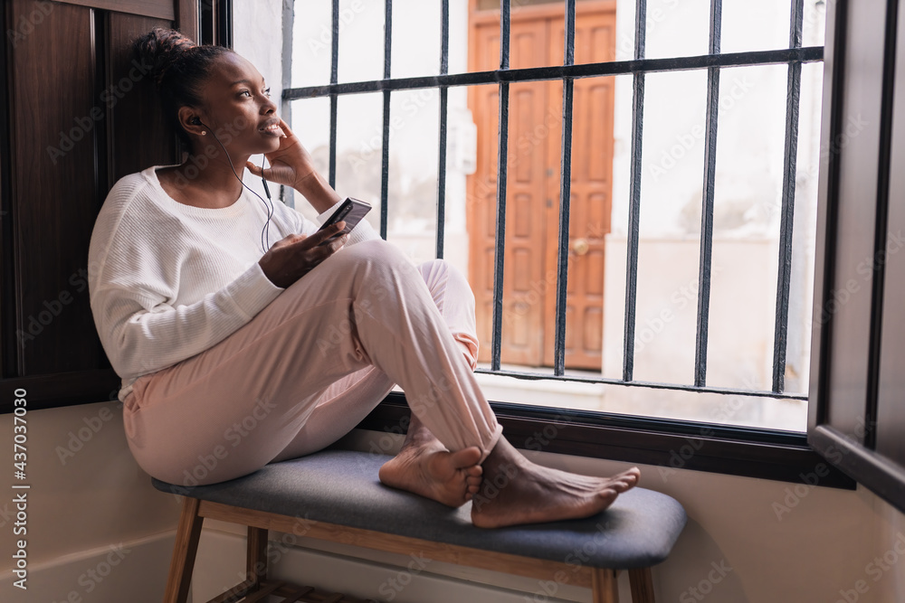 Young stylish black woman relaxing near window and enjoying music in earphones Young stylish black woman relaxing near window and enjoying music in earphones