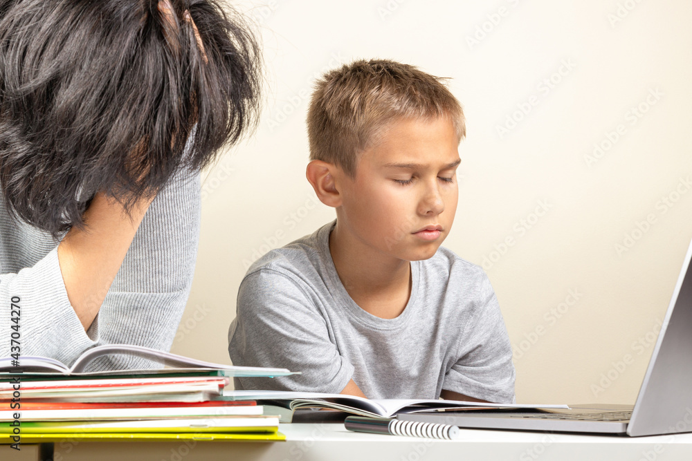 Disappointed frustrated tired exhausted sad kid sits at table with laptop computer and textbooks. Mother try to him help to do homework. Learning difficulties, education at home Disappointed frustrated tired exhausted sad kid sits at table with laptop computer and textbooks. Mother try to him help to do homework. Learning difficulties, education at home