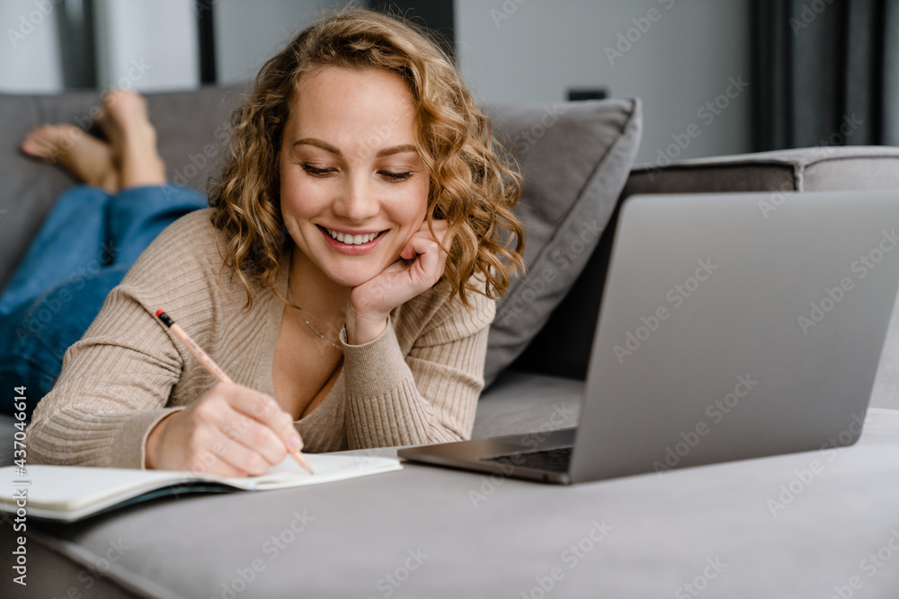 White woman writing down notes and using laptop while lying on couch White woman writing down notes and using laptop while lying on couch