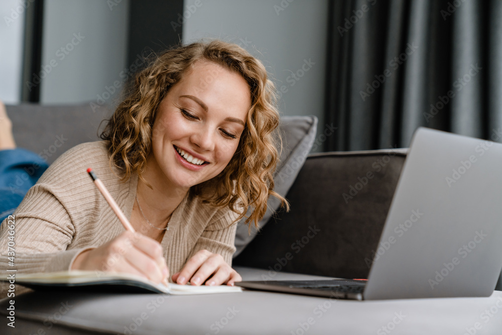 White woman writing down notes and using laptop while lying on couch White woman writing down notes and using laptop while lying on couch