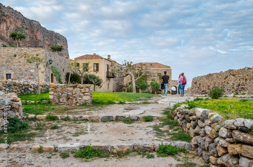 Tourists Visit Historic Castle Town in Monemvasia Island, Greece. Traditional Houses and Medieval City Walls in a Natural Environment with Trees and Gardens