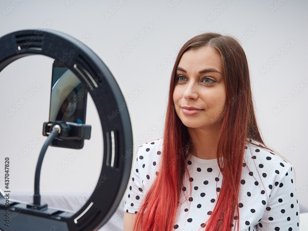 Beautiful woman blogger in a white t-shirt with polka dots, writes an online lesson, using a smartphone and a ring lamp Beautiful woman blogger in a white t-shirt with polka dots, writes an online lesson, using a smartphone and a ring lamp