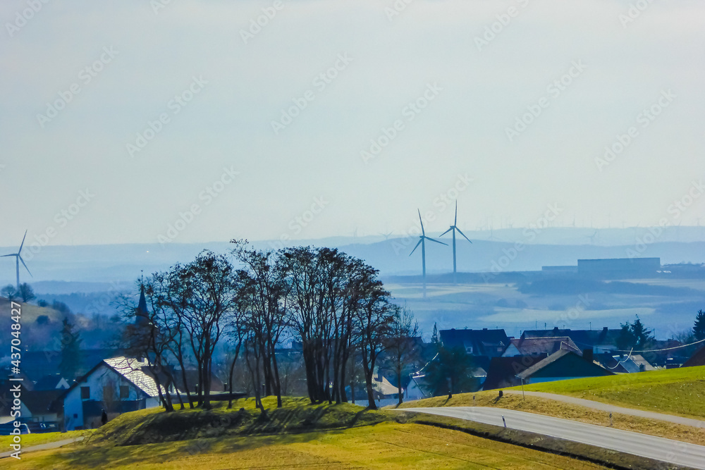 Hügelige Landschaft mit blauem Dunst, Windenergieanlagen, dunklen Bäumen, und gelben Feldern. Landscape in Eifel, Germany. Wind turbine and renewable energy in germany. Idyllisch concept renewables.