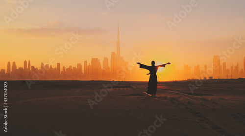 Arabic woman weared in traditional UAE dress - abayain rising her hands on the sunset at a desert with Dubai city silhouette on the background.