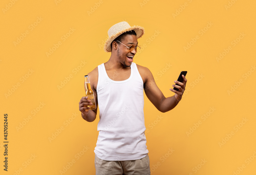 Young black guy with bottle of beer looking at his cellphone in excitement, winning lottery or casino, yellow background